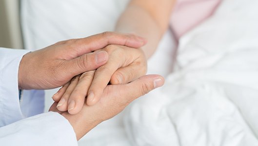 Patienten & Besucher: Die Hand einer im Bett liegenden Patientin wird von den beiden Händen einer Ärztin tröstend umfasst. Das Foto zeigt allein die Hände.